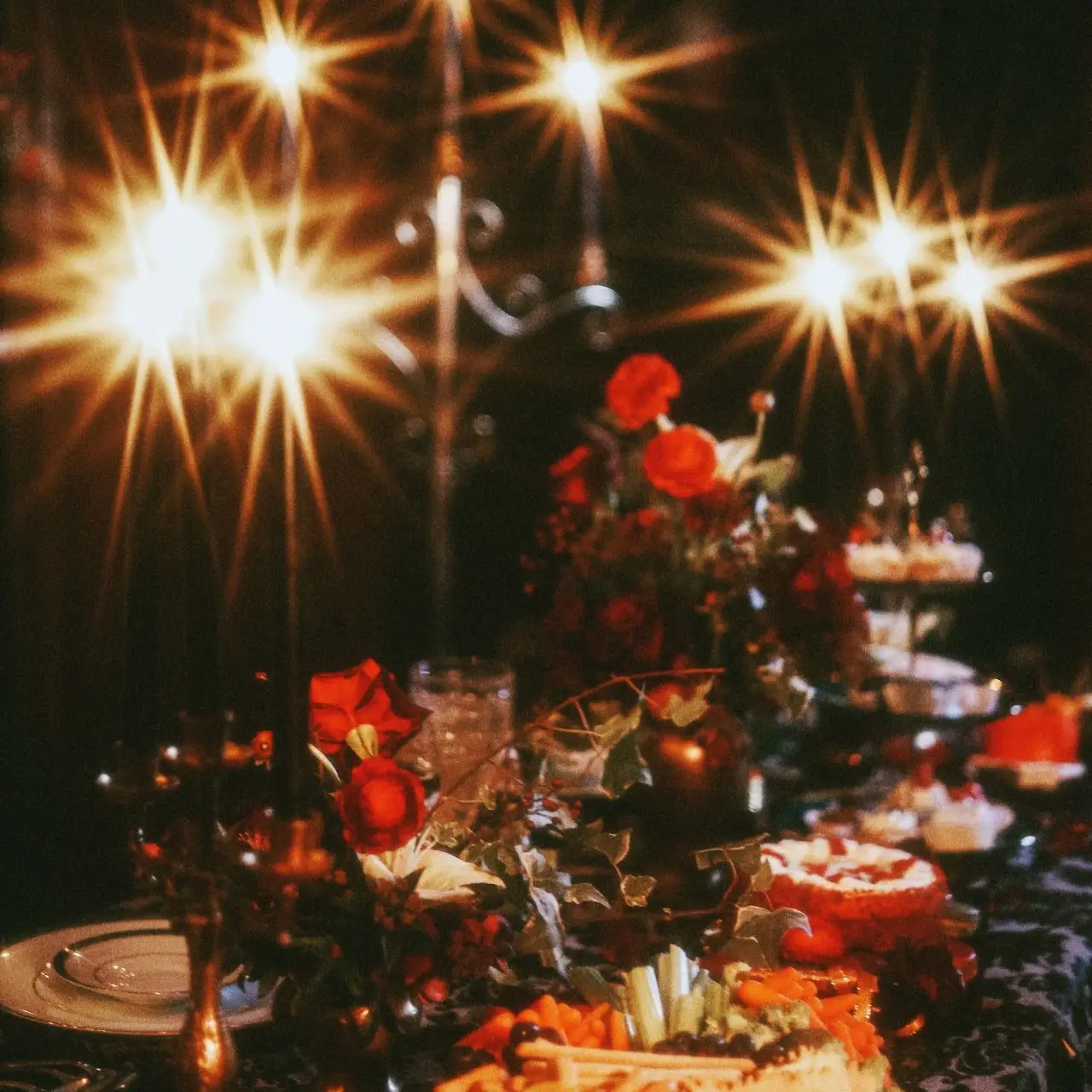 A candlelit tea table filled with red flowers, cakes and antique serving pieces